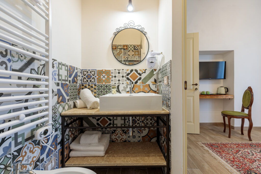 Bagno privato di Villa Fontis Rapolano con ampio lavabo d'appoggio, pareti rivestite in maioliche decorative, specchio rotondo, asciugamani e vista sulla camera matrimoniale, ideale per un soggiorno vicino alle Terme di Rapolano