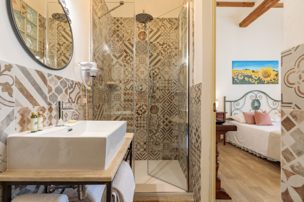 Bagno privato di Villa Fontis Rapolano con doccia walk-in in vetro, lavabo moderno e rivestimenti decorativi in gres; sullo sfondo si intravede la luminosa camera matrimoniale in stile toscano