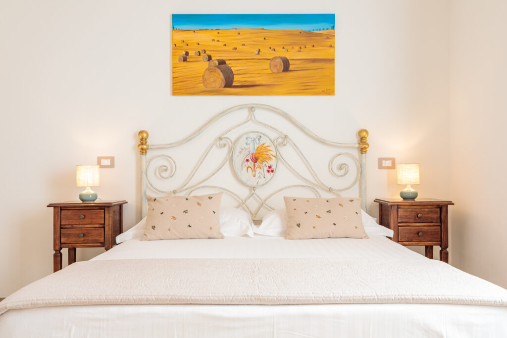 Camera matrimoniale di Villa Fontis Rapolano con elegante letto in ferro battuto decorato, comodini in legno, illuminazione soffusa e dipinto ispirato ai paesaggi della campagna toscana
