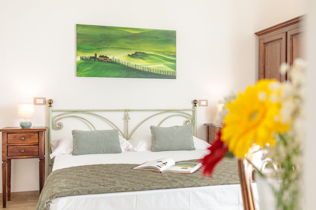 Camera matrimoniale di Villa Fontis Rapolano con letto matrimoniale, testiera in ferro battuto, comodini in legno, quadro paesaggistico toscano, lampade da comodino e fiori in primo piano.