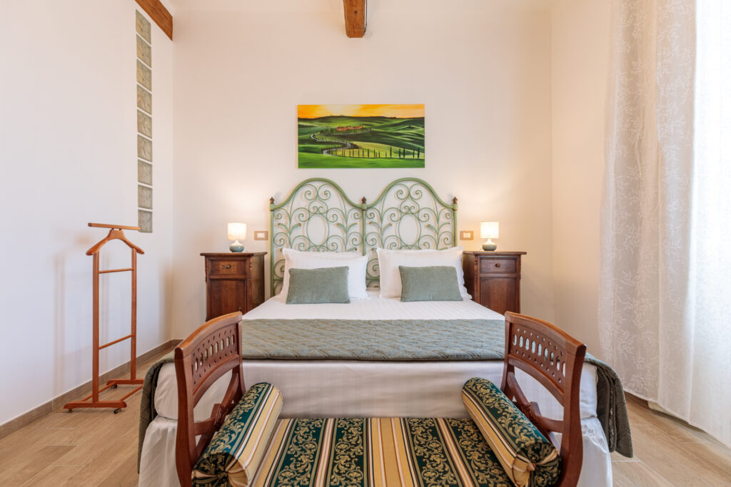 Camera matrimoniale di Villa Fontis Rapolano con letto king size, testiera in ferro battuto verde, comodini in legno, panca ai piedi del letto, travi a vista e arredi in autentico stile toscano.