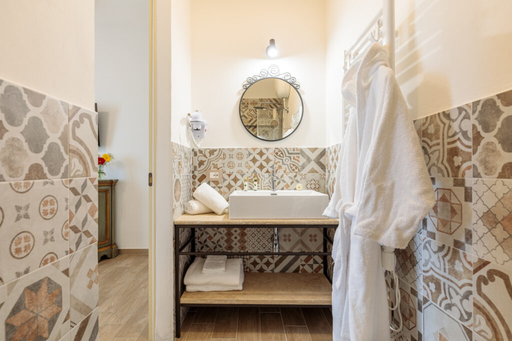 Bagno privato di Villa Fontis Rapolano con lavabo in pietra naturale, specchio rotondo, termoarredo, asciugamani, asciugacapelli e rivestimenti in maiolica decorata in stile toscano